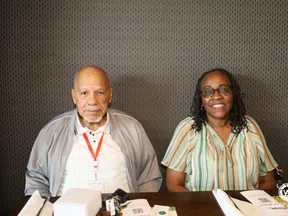 Smiling elderly couple sitting at a table with papers and personal items against patterned wall.