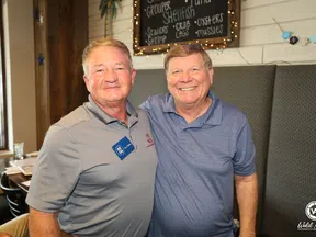 Two smiling middle-aged men standing together in a restaurant with seafood menu on chalkboard behind them