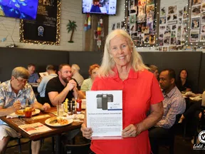 Smiling woman in red shirt holding informational flyer in busy casual restaurant with diners eating and socializing.