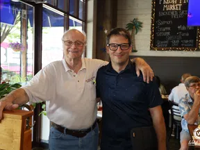 Two men smiling with arm around each other inside a casual restaurant with menu board in background