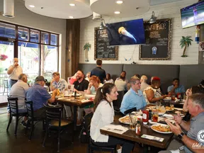Busy restaurant interior with diverse diners enjoying meals under TV screens and rustic decor on walls.