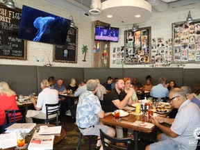 Busy seafood restaurant interior with customers dining and watching TVs, chalkboard menu on wall listing fish options