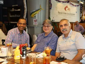 Three men smiling and seated at a restaurant table with drinks, condiments, and food at Hook Line & Schooner.