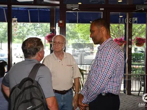 Three men engaged in conversation inside a well-lit cafe with glass doors and flower pots visible outside.