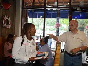 Woman in white shirt holding pamphlets conversing with elderly man in casual clothes inside a cafe.