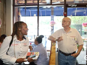Woman holding brochures talks to an elderly man inside a bright cafe with large windows and hanging plants.