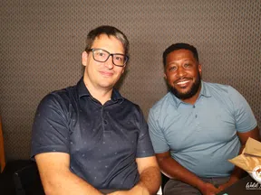 Two men smiling and sitting side by side against a patterned background, one wearing glasses and the other holding a paper bag.
