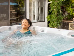 Woman relaxing in a bubbling outdoor hot tub near a house with greenery and patio furniture.