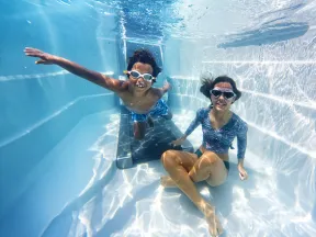 Two children wearing goggles underwater in a swimming pool, playing and smiling near a pool ladder.