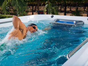 Man swimming vigorously in a small outdoor swim spa surrounded by greenery and a digital timer.