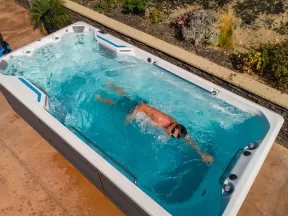 Man swimming in a blue swim spa with water jets in an outdoor patio setting on a sunny day