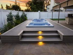Modern backyard with illuminated steps leading to a built-in hot tub on a wooden deck at sunset.