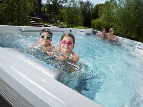 Two children with goggles and two adults relaxing in a backyard outdoor hot tub on a sunny day.