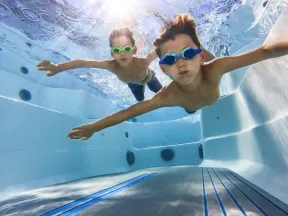 Two boys wearing swimming goggles underwater in a pool, swimming towards the camera with arms outstretched.