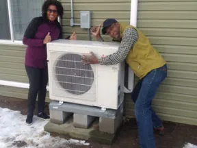 Two people giving thumbs up next to an outdoor HVAC unit mounted on a concrete base beside a green house in snowy yard.