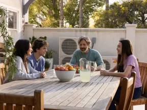 Family of four enjoying a sunny outdoor meal with lemonade and fruit on a wooden table in a backyard patio.