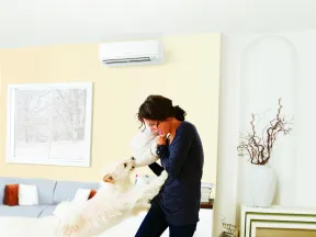Woman playing with her white fluffy dog on a couch in a bright, modern living room with neutral decor.