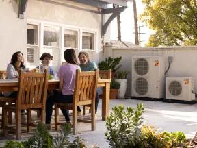 Family of four sitting at an outdoor wooden table enjoying a meal on a sunny patio with air conditioning units nearby