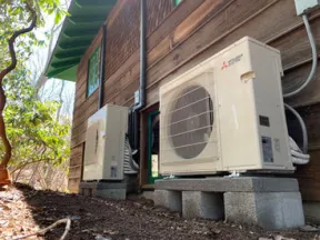 Two Mitsubishi outdoor air conditioning units installed on concrete blocks outside a wooden house surrounded by trees.