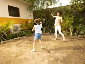 Woman and girl playing badminton outside in sunny backyard near yellow house and green plants.