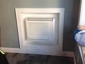 Wall-mounted air conditioning unit with white decorative trim on gray wall above tiled floor.