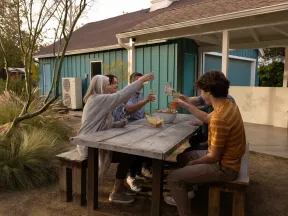 Group of friends sitting outdoors at a rustic wooden table toasting drinks and enjoying a sunny day.