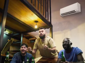 Three men excitedly watching a sports game indoors with snacks and drinks on the table and an air conditioner on the wall.