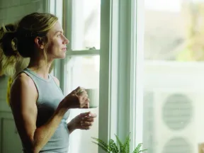 Woman holding a cup, looking out a bright window in a cozy indoor setting with plants nearby.