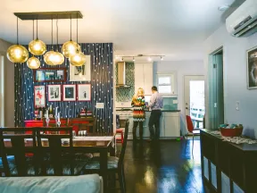 Modern dining and kitchen area with hanging globe lights, patterned wall, wooden dining set, and two people near the stove.