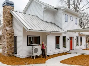 Modern two-story white house with metal roof, stone chimney, red door, and man standing by outdoor HVAC unit.