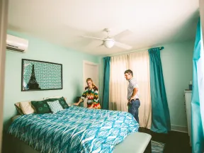 Couple examines bed with blue patterned bedding in a light turquoise bedroom with matching curtains and wall art.