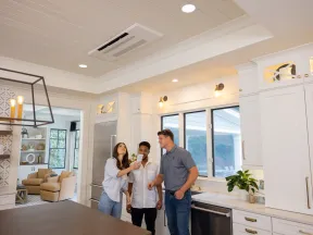Three friends smiling and taking a selfie in a bright modern kitchen with white cabinets and large windows.