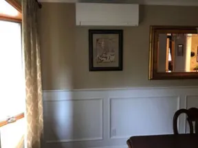 Cozy dining room corner with wooden table, patterned rug, wall art, large mirror, window with curtains, and air conditioner unit.