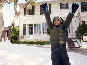 Happy child in winter clothes plays in fresh snow in front of a white house with an American flag.