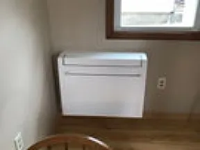 Wooden chair in front of a white heater mounted on a beige wall under a window with sheer curtains.