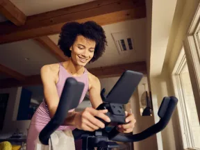 Smiling woman in pink workout clothes using stationary bike in sunlit room with wooden beams and large windows.