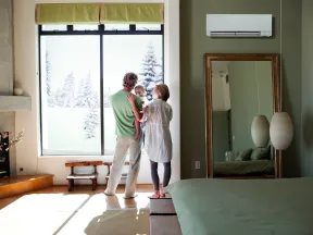 A family of three stands by a large window in a cozy bedroom, looking out at snow-covered trees during winter.