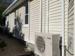Exterior beige house wall with a mounted Mitsubishi outdoor air conditioning unit and white window frames.