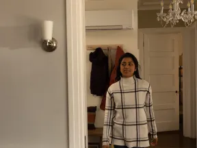 Young woman in a plaid sweater walking through a doorway into a cozy home with coats and chandelier visible.