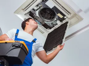 Technician in blue overalls inspecting and cleaning a ceiling air conditioning unit while holding a toolbox on a ladder.