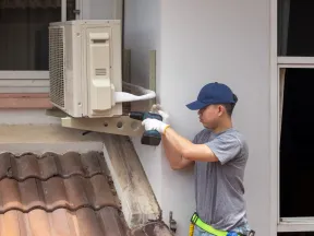 Technician wearing gloves and cap installs an air conditioning unit on a house exterior wall above a tiled roof.