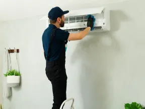Technician in uniform servicing a wall-mounted air conditioner unit on a gray wall inside a living room.