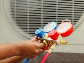 Technician's hand adjusting HVAC manifold gauge with blue and red hoses near an air conditioning unit outdoors.