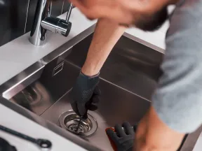 Plumber wearing gloves fixing a stainless steel kitchen sink drain using a tool and cloth