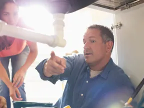 Plumber explaining repair work under a kitchen sink to a woman observing closely during maintenance.