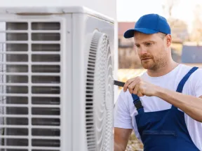 Technician in blue overalls and cap repairing an outdoor air conditioning unit with a screwdriver.