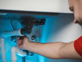 Plumber fixing white PVC pipes under a sink illuminated by blue light in a modern kitchen or bathroom.