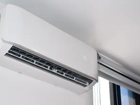 White wall-mounted air conditioner unit showing temperature set to 25 degrees near window with blinds.