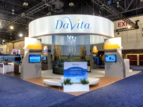 DaVita exhibition booth at a trade show with branded signage, digital screens, and seating area on blue carpet.