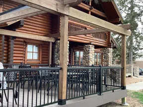 Log cabin porch with wooden beams, stone pillars, black metal railing, and outdoor seating under trees.